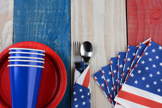 red plastic plate, blue plastic cups, silverware wrapped in a American flap paper napkin sitting on a red white blue stiped table 